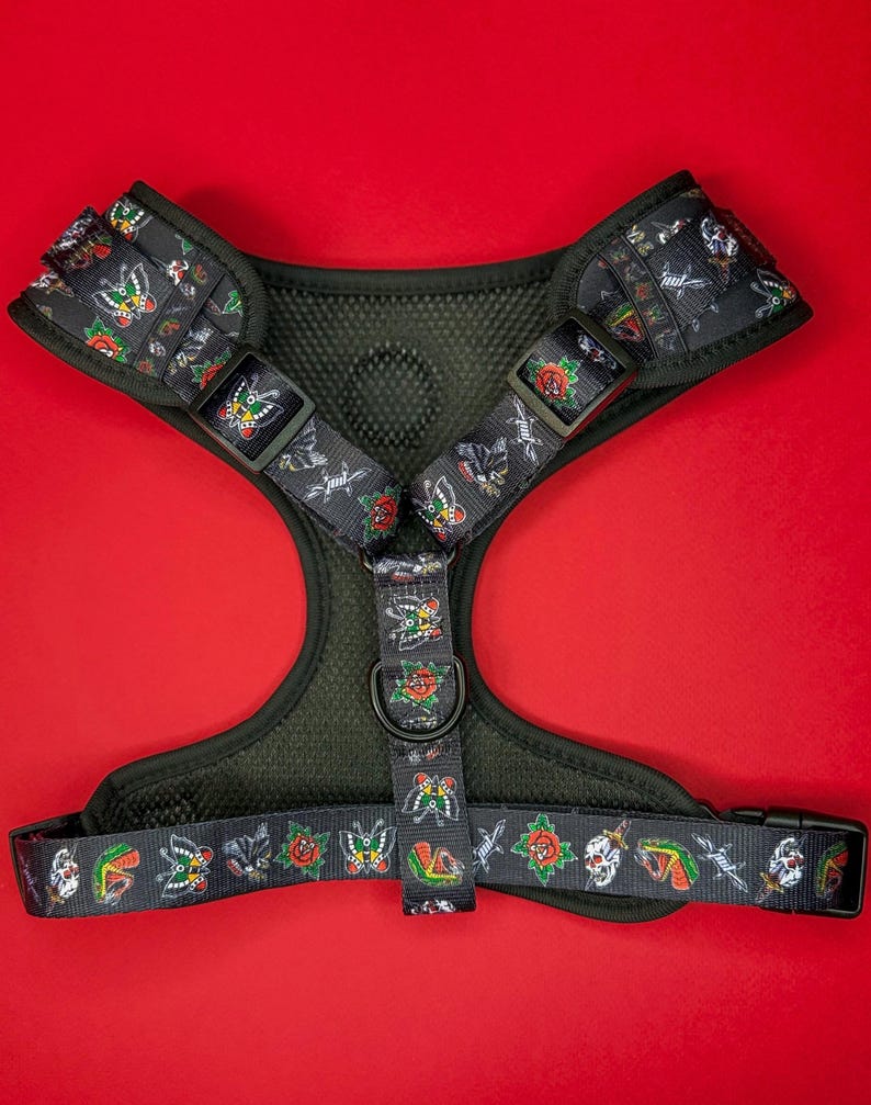 May include: A black dog harness with a colorful tattoo-inspired print featuring butterflies, roses, and skulls. The harness has a mesh lining and an adjustable buckle.