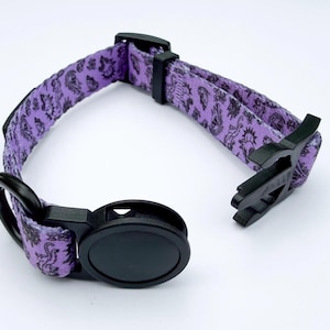May include: A purple pet collar featuring a repeating black graphic print of various small creatures. The collar includes a black plastic side-release buckle, a black plastic adjustment slider, and a black metal D-ring for attaching a leash.