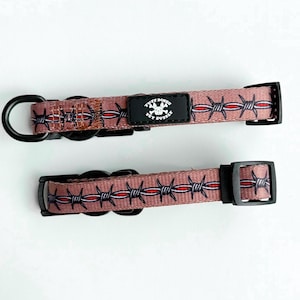 May include: Two dog collars with a barbed wire pattern in shades of pink, black, and red. The collars have black hardware, including a D-ring and a buckle. A black patch with white text and a skull logo is centered on the collar.