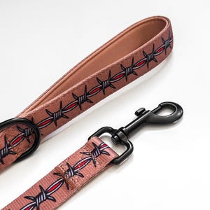 May include: A tan dog leash featuring a repeating barbed wire pattern with grey and red accents. The leash includes a black metal swivel snap hook and a black metal D-ring, with a looped handle design made from durable woven fabric material.