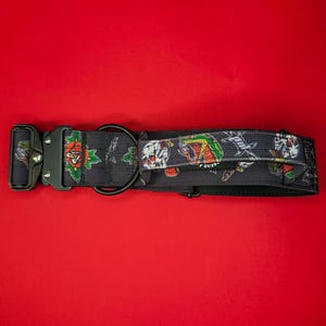May include: A black nylon dog collar featuring a repeating tattoo-style print of skulls, roses, and daggers. The collar includes a heavy-duty black metal side-release buckle, a black D-ring for leash attachment, and a small black fabric handle.
