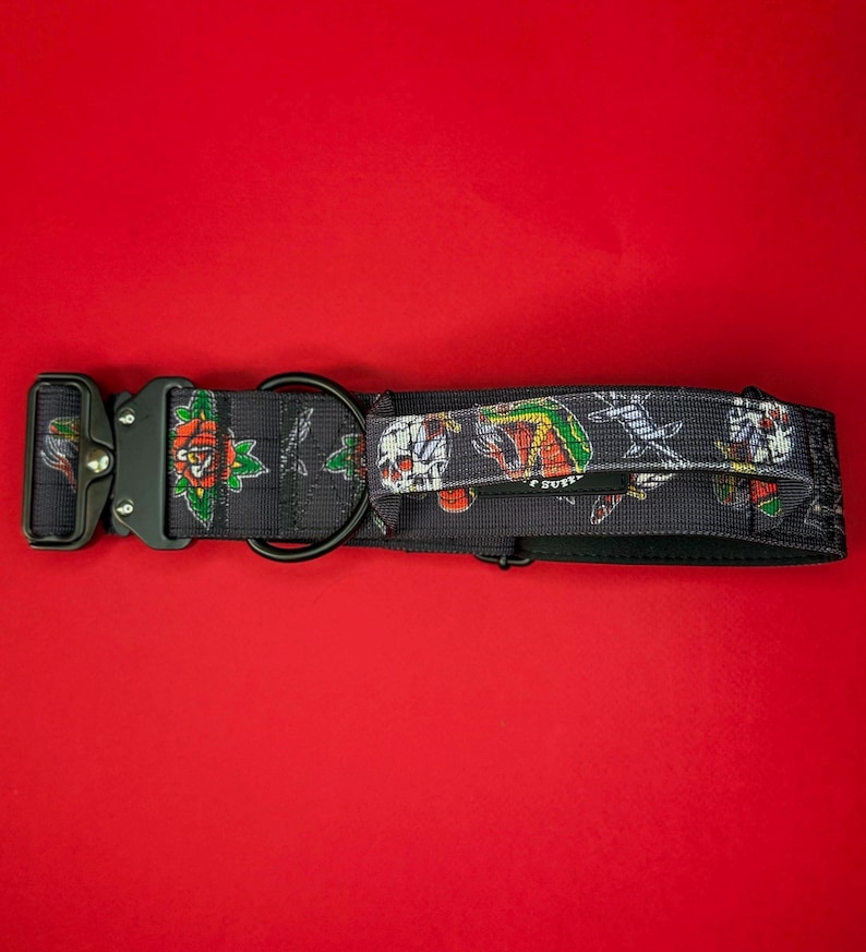 May include: A black nylon dog collar featuring a repeating tattoo-style print of skulls, roses, and daggers. The collar includes a heavy-duty black metal side-release buckle, a black D-ring for leash attachment, and a small black fabric handle.