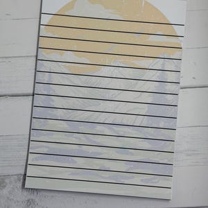 May include: A rectangular notepad featuring a landscape illustration with a large pale yellow sun, light blue mountains, and evergreen trees. The paper has horizontal black lines printed across the entire surface for writing notes or lists.