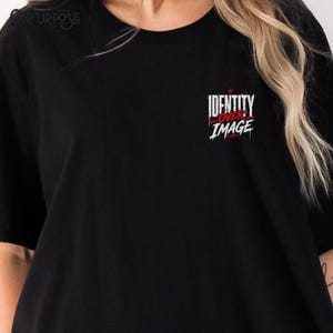 Pu&ograve; includere: Una t-shirt nera a maniche corte con una grafica sulla parte superiore sinistra del petto. La grafica mostra il testo bianco "IDENTITY" sopra il testo rosso "OVER" e il testo bianco "IMAGE" in un carattere stilizzato e usurato con una piccola croce rossa.