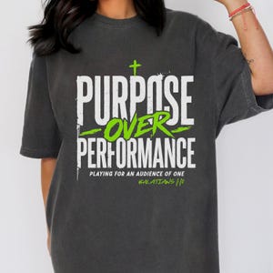 Può includere: Una t-shirt a maniche corte grigio antracite con testo bianco e verde lime che recita "PURPOSE OVER PERFORMANCE" con una piccola croce sopra, e il testo "PLAYING FOR AN AUDIENCE OF ONE" e "GALATIANS 1:10" stampato sotto.