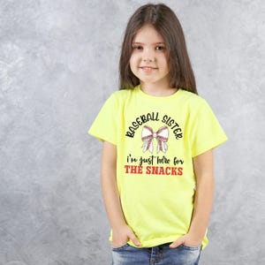 Graphic featuring the phrase 'Baseball Sister' in bold, curved lettering around a large white bow with red baseball stitching. Below, the text reads 'I'm just here for the snacks' in playful script, with 'THE SNACKS'