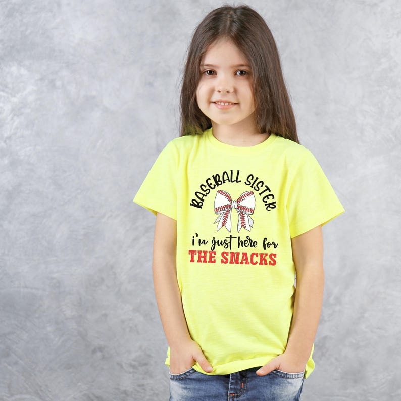 Graphic featuring the phrase 'Baseball Sister' in bold, curved lettering around a large white bow with red baseball stitching. Below, the text reads 'I'm just here for the snacks' in playful script, with 'THE SNACKS'