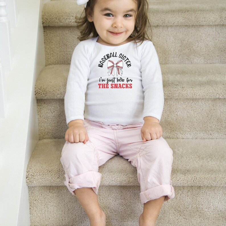 Graphic featuring the phrase 'Baseball Sister' in bold, curved lettering around a large white bow with red baseball stitching. Below, the text reads 'I'm just here for the snacks' in playful script, with 'THE SNACKS'