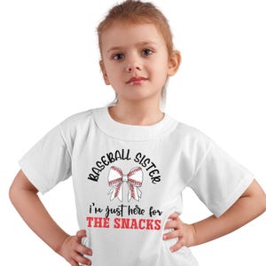 Graphic featuring the phrase 'Baseball Sister' in bold, curved lettering around a large white bow with red baseball stitching. Below, the text reads 'I'm just here for the snacks' in playful script, with 'THE SNACKS'