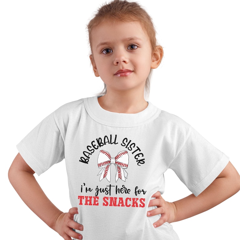 Graphic featuring the phrase 'Baseball Sister' in bold, curved lettering around a large white bow with red baseball stitching. Below, the text reads 'I'm just here for the snacks' in playful script, with 'THE SNACKS'