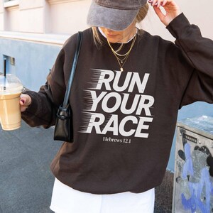 Può includere: Una felpa marrone scuro con la scritta bianca "RUN YOUR RACE" con effetto velocità. Il testo "Hebrews 12:1" è in basso. La felpa è indossata con un berretto da baseball e collane d'oro.