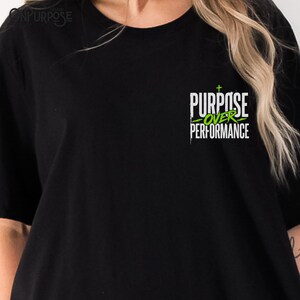 May include: A black short-sleeved t-shirt featuring a graphic on the upper left chest. The design includes the white text "PURPOSE" above the green text "OVER" and the white text "PERFORMANCE," with a small green cross above the word "PURPOSE."