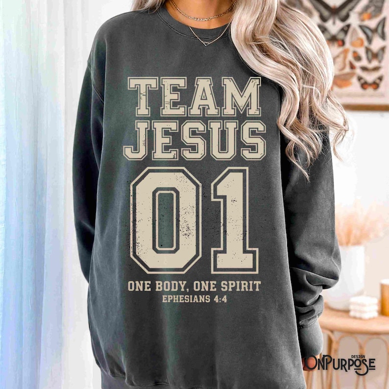 "Team Jesus 01" graphic design featuring bold varsity-style lettering in beige on a black background, with the Bible verse "One Body, One Spirit – Ephesians 4:4" displayed beneath. The design combines a sporty aesthetic with Christian faith messaging.