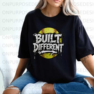 Pode incluir: Camiseta preta com a frase "BUILT DIFFERENT" em branco, sobre um gráfico de softball amarelo. O design inclui uma seta branca e um efeito de tinta escorrendo. A camiseta é de estilo casual.