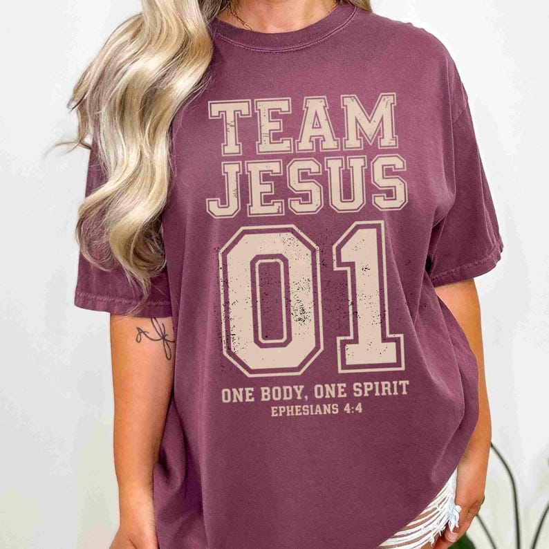"Team Jesus 01" graphic design featuring bold varsity-style lettering in beige on a black background, with the Bible verse "One Body, One Spirit – Ephesians 4:4" displayed beneath. The design combines a sporty aesthetic with Christian faith messaging.