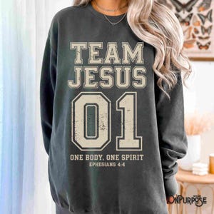 "Team Jesus 01" graphic design featuring bold varsity-style lettering in beige on a black background, with the Bible verse "One Body, One Spirit – Ephesians 4:4" displayed beneath. The design combines a sporty aesthetic with Christian faith messaging.