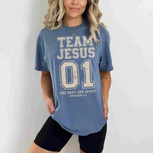 "Team Jesus 01" graphic design featuring bold varsity-style lettering in beige on a black background, with the Bible verse "One Body, One Spirit – Ephesians 4:4" displayed beneath. The design combines a sporty aesthetic with Christian faith messaging.