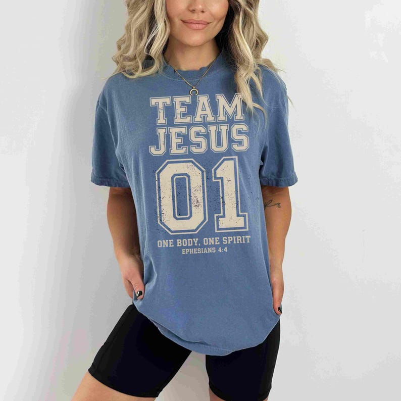 "Team Jesus 01" graphic design featuring bold varsity-style lettering in beige on a black background, with the Bible verse "One Body, One Spirit – Ephesians 4:4" displayed beneath. The design combines a sporty aesthetic with Christian faith messaging.