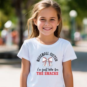 Graphic featuring the phrase 'Baseball Sister' in bold, curved lettering around a large white bow with red baseball stitching. Below, the text reads 'I'm just here for the snacks' in playful script, with 'THE SNACKS'