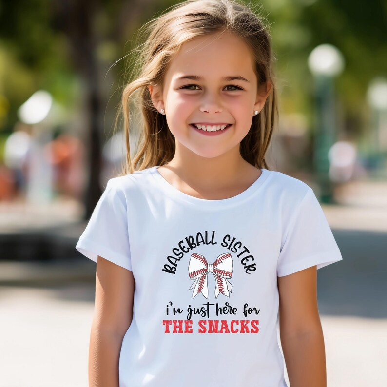 Graphic featuring the phrase 'Baseball Sister' in bold, curved lettering around a large white bow with red baseball stitching. Below, the text reads 'I'm just here for the snacks' in playful script, with 'THE SNACKS'