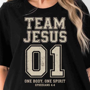 "Team Jesus 01" graphic design featuring bold varsity-style lettering in beige on a black background, with the Bible verse "One Body, One Spirit – Ephesians 4:4" displayed beneath. The design combines a sporty aesthetic with Christian faith messaging.