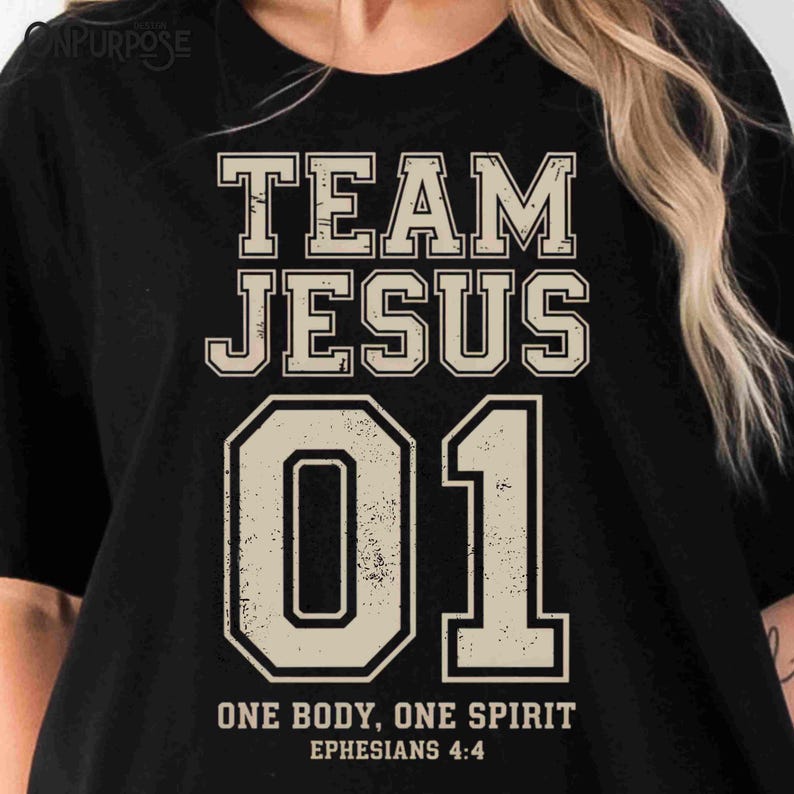 "Team Jesus 01" graphic design featuring bold varsity-style lettering in beige on a black background, with the Bible verse "One Body, One Spirit – Ephesians 4:4" displayed beneath. The design combines a sporty aesthetic with Christian faith messaging.