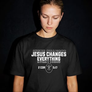 May include: Black t-shirt featuring the white text "JESUS CHANGES EVERYTHING". Underneath is "2 COR 5:17" with a cross and circle graphic. The shirt is worn by a person.