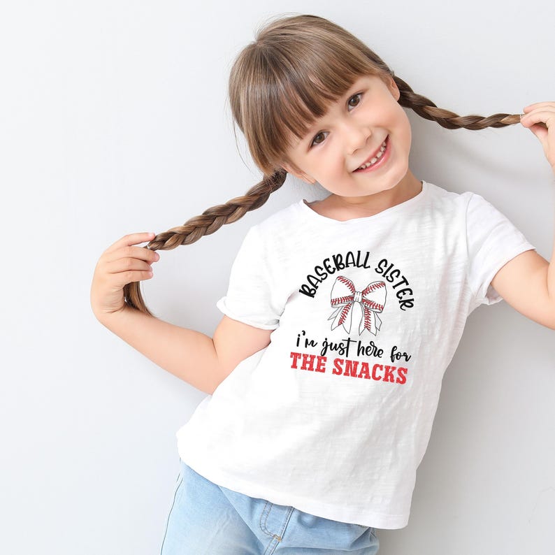 Graphic featuring the phrase 'Baseball Sister' in bold, curved lettering around a large white bow with red baseball stitching. Below, the text reads 'I'm just here for the snacks' in playful script, with 'THE SNACKS'