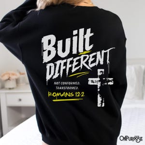 Puede incluir: Una sudadera negra de manga larga con texto gráfico blanco y amarillo en la espalda que dice Built Different, Not Conformed. Transformed. Romans 12:2, acompañado de un gráfico de cruz blanca con una textura desgastada.