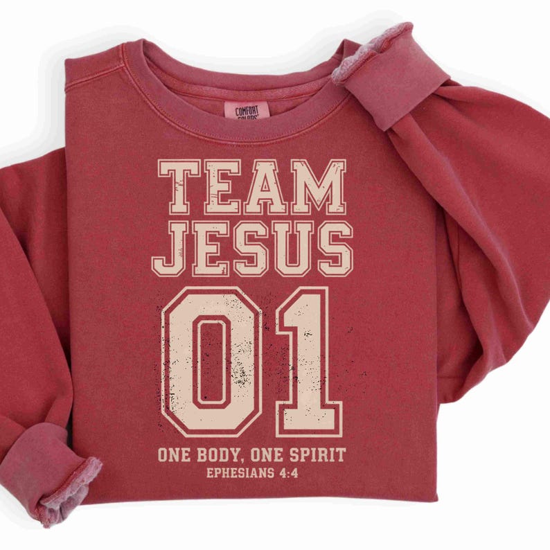 "Team Jesus 01" graphic design featuring bold varsity-style lettering in beige on a black background, with the Bible verse "One Body, One Spirit – Ephesians 4:4" displayed beneath. The design combines a sporty aesthetic with Christian faith messaging.