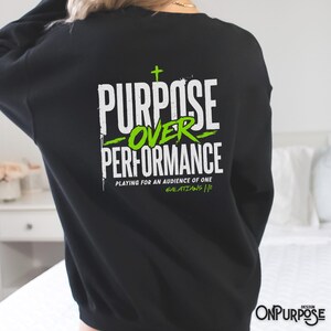 May include: A black long-sleeve sweatshirt featuring a white and lime green graphic on the back. The text reads PURPOSE OVER PERFORMANCE, PLAYING FOR AN AUDIENCE OF ONE, and GALATIANS 1:10, with a small cross symbol above the word PURPOSE.