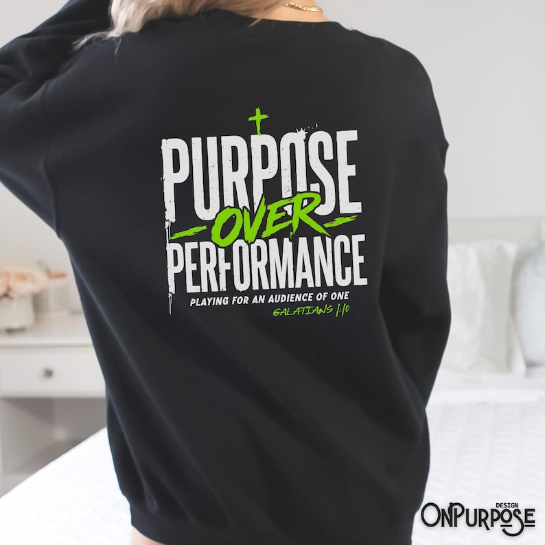 May include: A black long-sleeve sweatshirt featuring a white and lime green graphic on the back. The text reads PURPOSE OVER PERFORMANCE, PLAYING FOR AN AUDIENCE OF ONE, and GALATIANS 1:10, with a small cross symbol above the word PURPOSE.