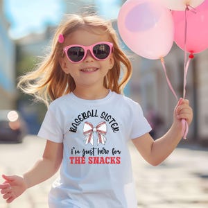Graphic featuring the phrase 'Baseball Sister' in bold, curved lettering around a large white bow with red baseball stitching. Below, the text reads 'I'm just here for the snacks' in playful script, with 'THE SNACKS'