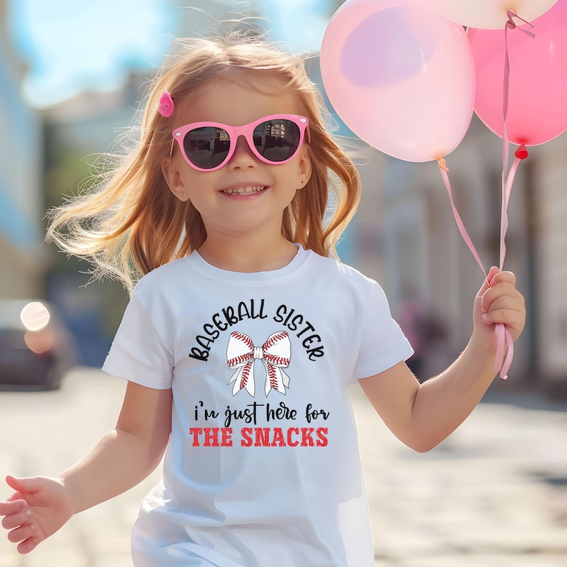 Graphic featuring the phrase 'Baseball Sister' in bold, curved lettering around a large white bow with red baseball stitching. Below, the text reads 'I'm just here for the snacks' in playful script, with 'THE SNACKS'