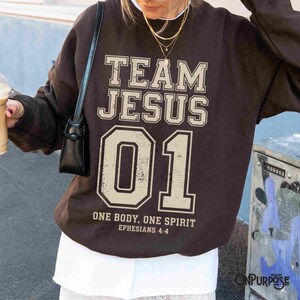 "Team Jesus 01" graphic design featuring bold varsity-style lettering in beige on a black background, with the Bible verse "One Body, One Spirit – Ephesians 4:4" displayed beneath. The design combines a sporty aesthetic with Christian faith messaging.
