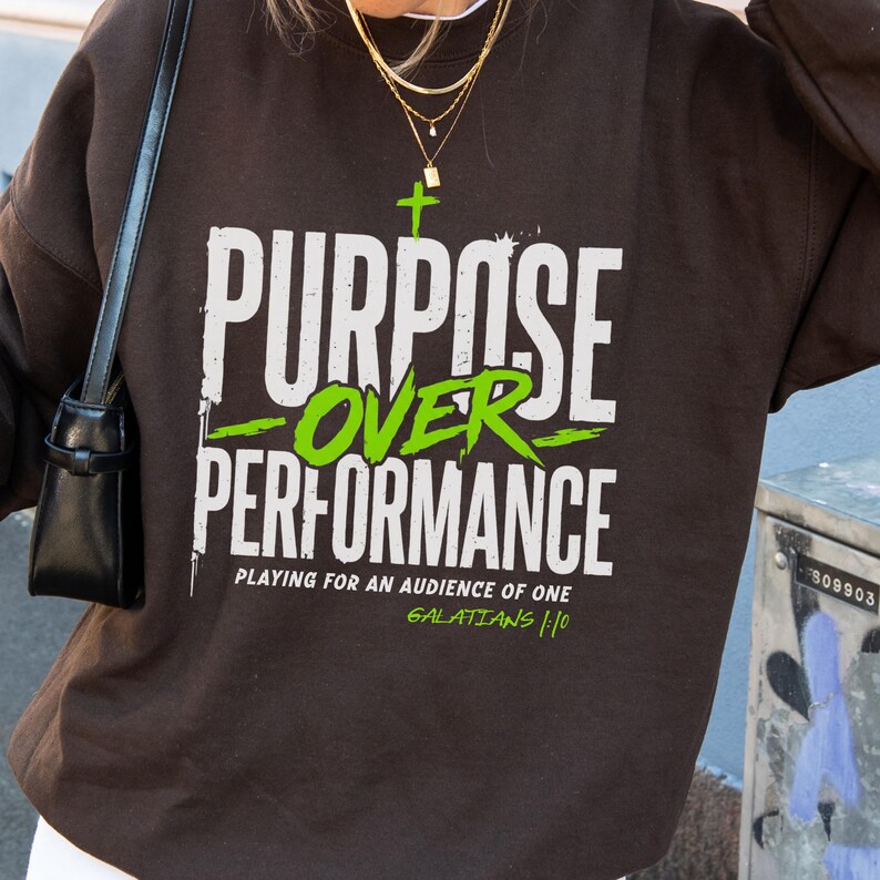 May include: A dark brown long-sleeve sweatshirt featuring white and lime green text that reads "PURPOSE OVER PERFORMANCE" with a small cross above the text. Below, it says "PLAYING FOR AN AUDIENCE OF ONE" and "GALATIANS 1:10" in lime green.