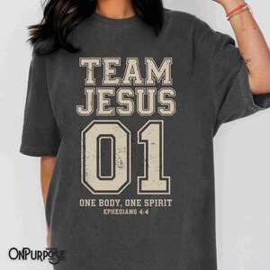 "Team Jesus 01" graphic design featuring bold varsity-style lettering in beige on a black background, with the Bible verse "One Body, One Spirit – Ephesians 4:4" displayed beneath. The design combines a sporty aesthetic with Christian faith messaging.