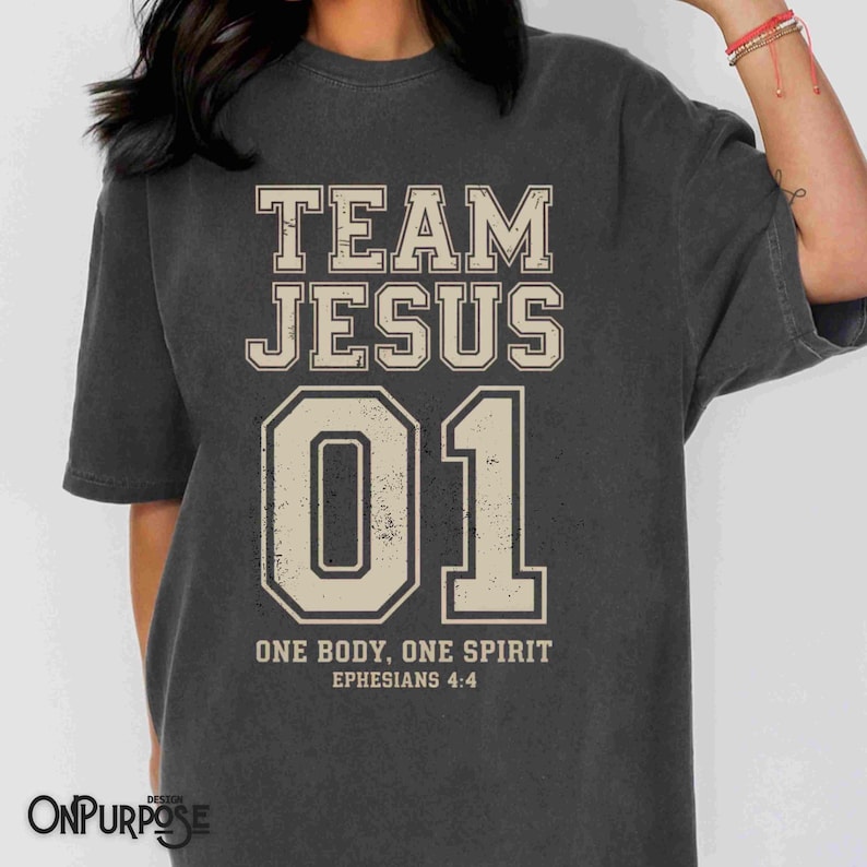 "Team Jesus 01" graphic design featuring bold varsity-style lettering in beige on a black background, with the Bible verse "One Body, One Spirit – Ephesians 4:4" displayed beneath. The design combines a sporty aesthetic with Christian faith messaging.