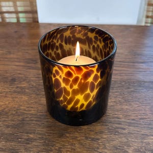 May include: A lit candle in a brown and amber glass votive holder. The glass has a spotted pattern. The candle is white and the flame is yellow. The votive holder is sitting on a wooden surface.