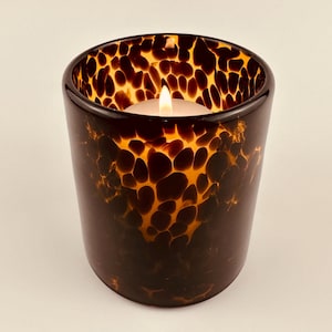 May include: A cylindrical candle holder with a brown and amber spotted pattern. A small white candle with a lit flame is inside the holder. The glass has a glossy finish. The candle holder is a decorative item.