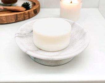 May include: A white round bar of soap sits on a gray and white marble soap dish. The dish has a raised rim and a flat base.