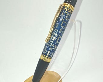 May include: A stylish pen with a black top and bottom, gold accents, and a blue barrel featuring a circuit board design. The pen is displayed on a small, gold-colored stand.