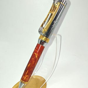May include: A decorative pen with a silver and gold metal design, featuring a skull on the top. The pen has a red and gold marbled barrel. The pen is displayed on a clear stand.