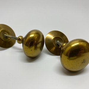 May include: Two antique brass door knobs with a golden finish. Each knob has a round, bulbous shape and is connected to a circular backplate by a slender rod. The knobs are likely vintage and add a classic touch.