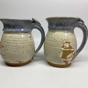 May include: Two handmade ceramic mugs. The mugs have a speckled beige body with a blue rim and handle. One mug has a brown design on the side. The mugs are likely used for drinking beverages.