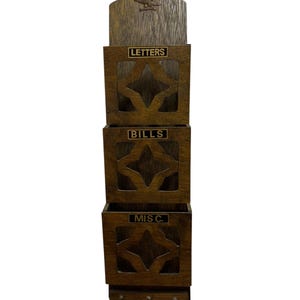 May include: A vertical wooden wall organiser with three stacked compartments labelled LETTERS, BILLS, and MIS C. in black text. The unit features a dark brown wood grain finish, a carved eagle emblem at the top, and three small metal hooks at the bottom.
