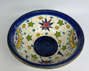 May include: Hand-painted ceramic bowl with a blue rim and a brass-coloured edge. The interior features a cream-coloured base adorned with colourful stars and sun-like designs. The centre of the bowl is a deep blue.