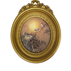 May include: An oval-shaped, gold-colored picture frame with a detailed, ornate design. The framed artwork depicts two birds perched on a branch, with a nest and a sunset in the background. The frame measures approximately 8 inches in diameter.