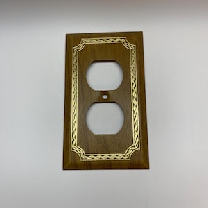 May include: A brown wooden outlet cover with a gold decorative border. The cover has a single outlet.