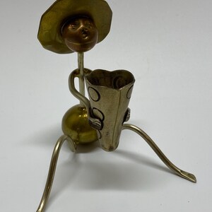 May include: A gold metal figurine of a person wearing a hat and holding a cup. The figurine has a round body and long, thin legs. The cup has a decorative design.